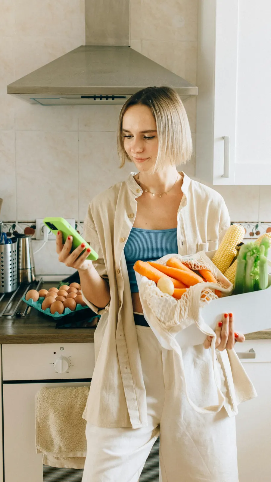 Une femme commande sur son téléphone en tenant un sac de légumes frais dans sa cuisine